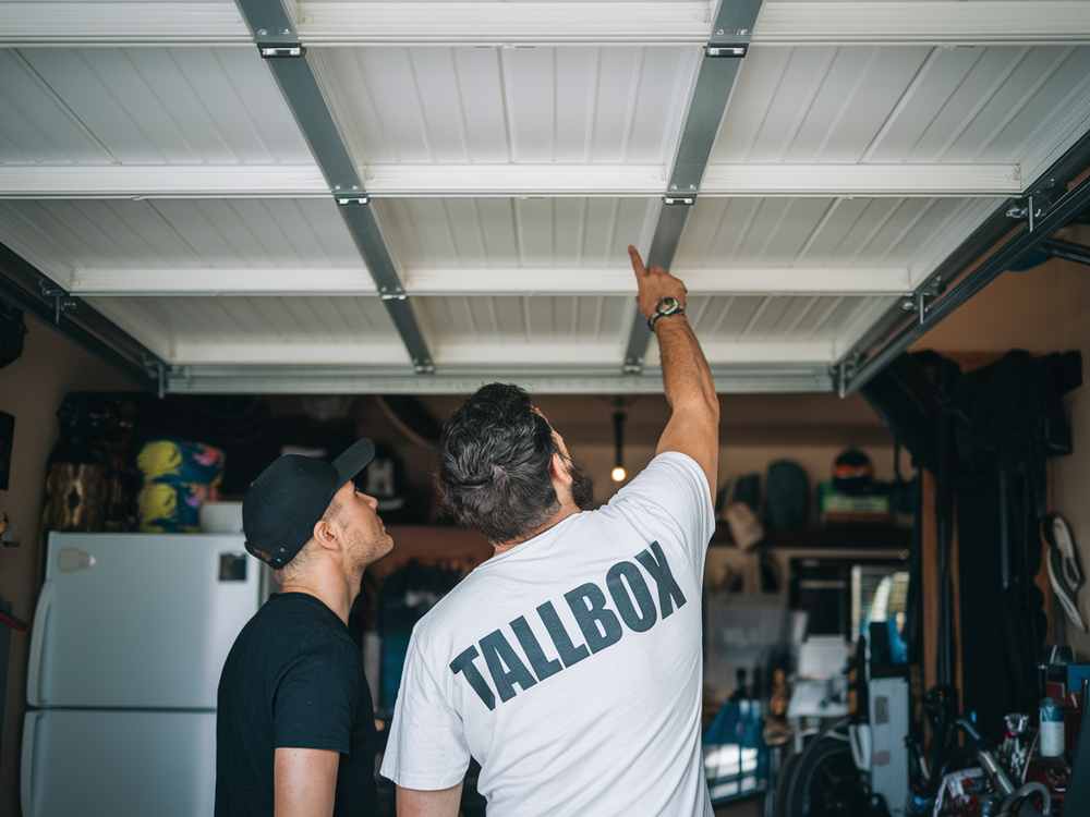 Two men in a garage, one pointing at the ceiling, discussing garage door maintenance tips. A fridge and various items are visible in the background.