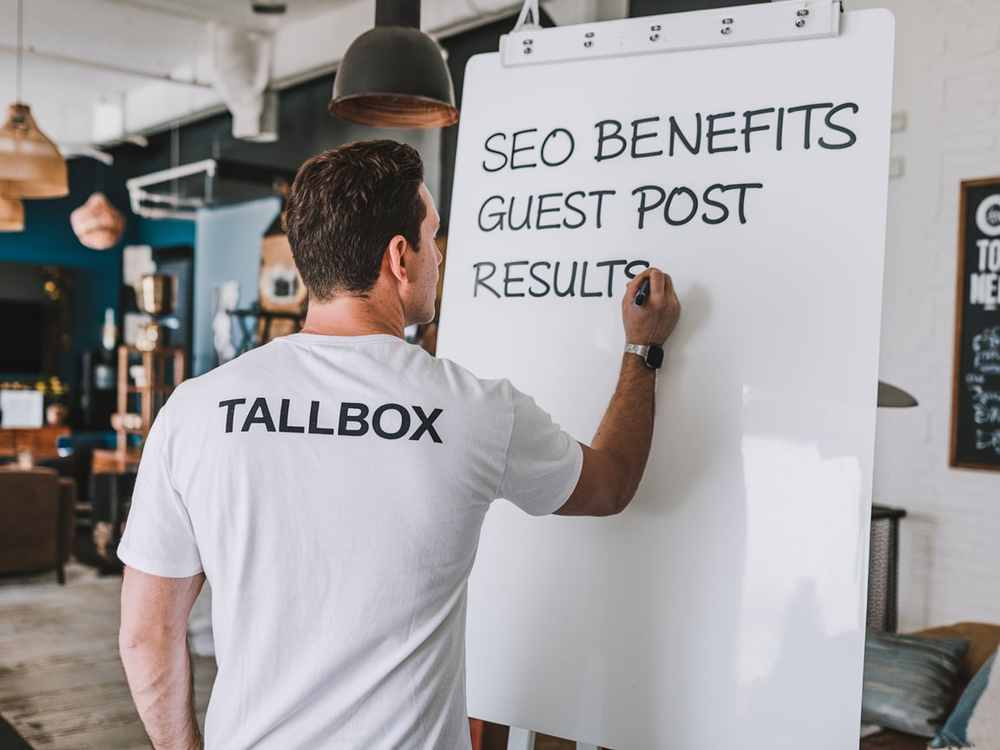 In an office, a man writes "SEO benefits, guest post, results" on a whiteboard, illustrating how guest blogging can enhance visibility and credibility.