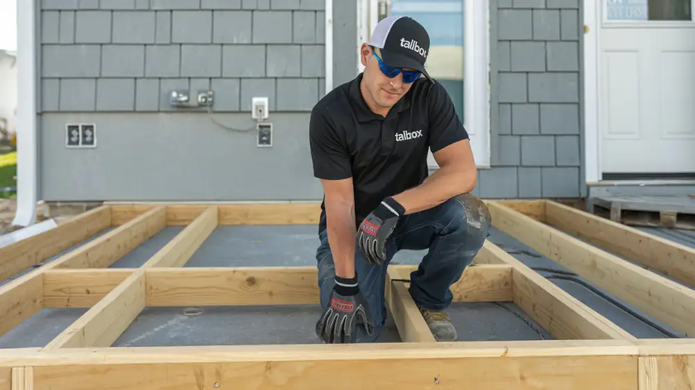 A person in a black shirt and cap is kneeling, diligently working on the wooden frame of an outdoor kitchen on a house deck.