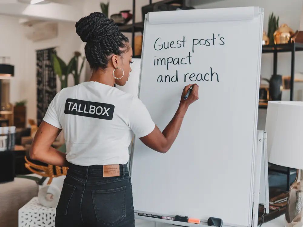 A person in a "TALLBOX" shirt writes on a flip chart that reads "Guest post’s impact and reach," highlighting the power of guest blogging for interior designers looking to boost their SEO strategies.