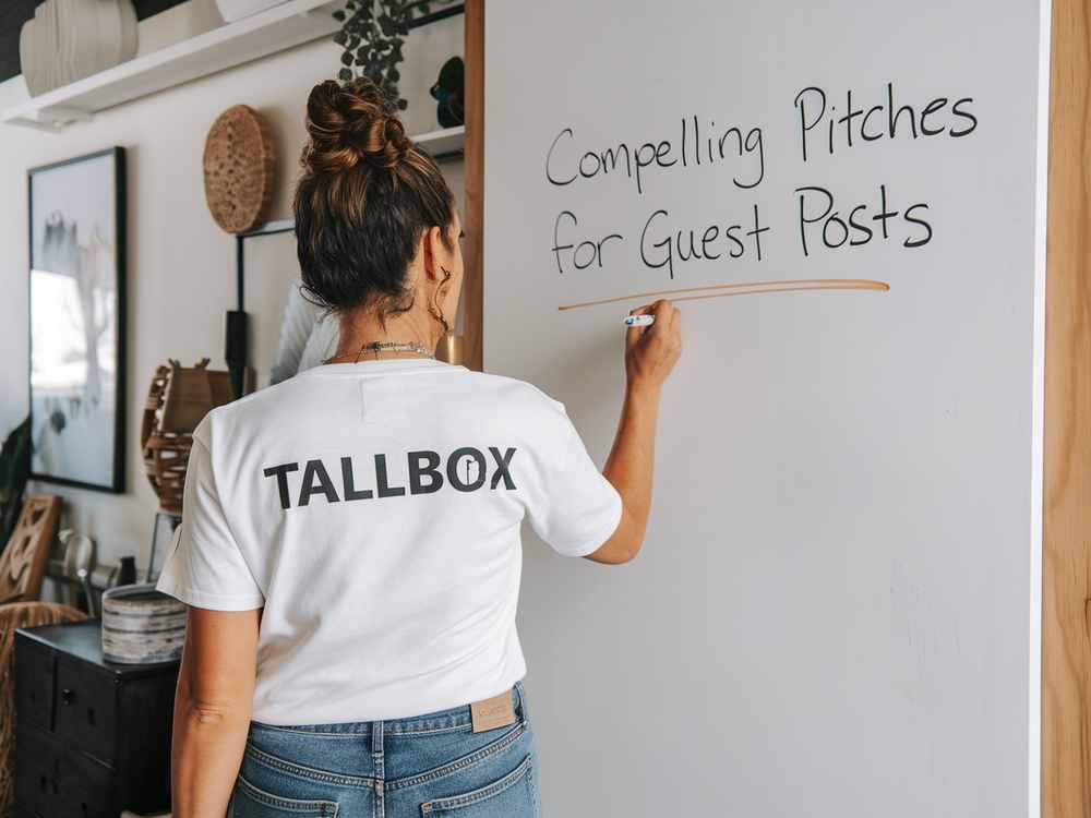 In an office setting, a woman wearing a "Tallbox" shirt scribbles "Compelling Pitches for Guest Posts" on the whiteboard, focusing on strategies that capture the attention of real estate agents and enhance guest blogging efforts for optimal SEO results.