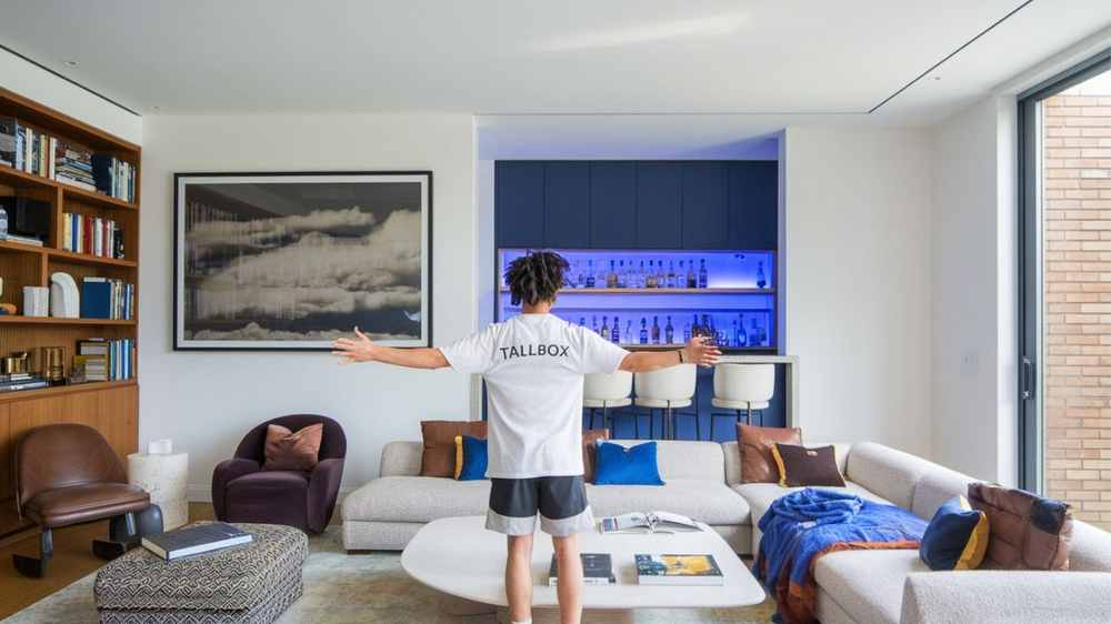 Person with arms outstretched in a modern living room, showcasing luxury renovations like the bespoke bookshelves and a chic blue-lit bar area, highlighting where to spend and save for an elegant yet practical transformation.