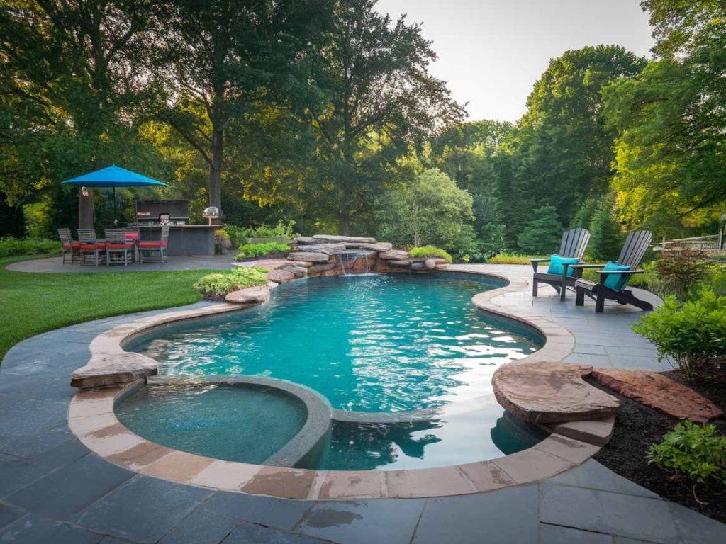Experience the ultimate in outdoor living with this luxury pool, featuring a curved design and a soothing waterfall. Surrounded by a patio, comfortable chairs, and vibrant trees, it offers a serene oasis in your lush garden setting.