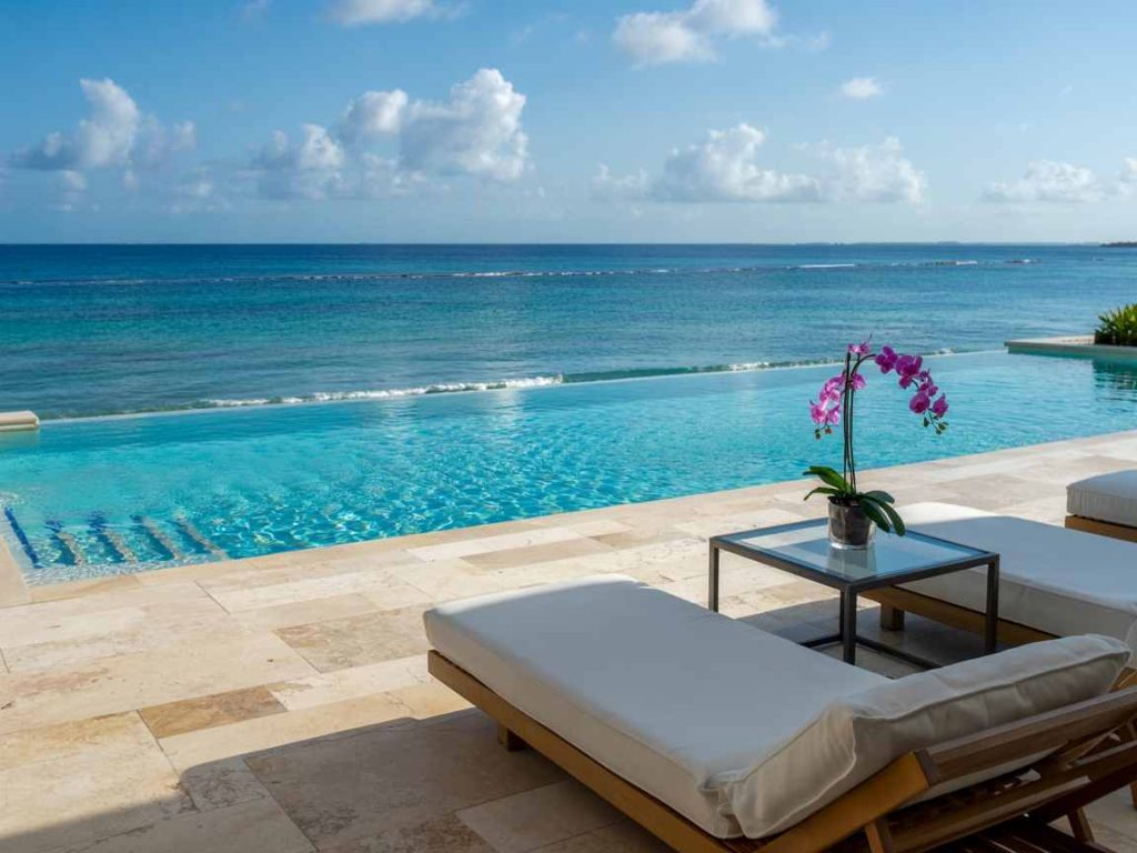 Lounge chairs and a table with an orchid beside a luxury pool overlooking the ocean under a blue sky.