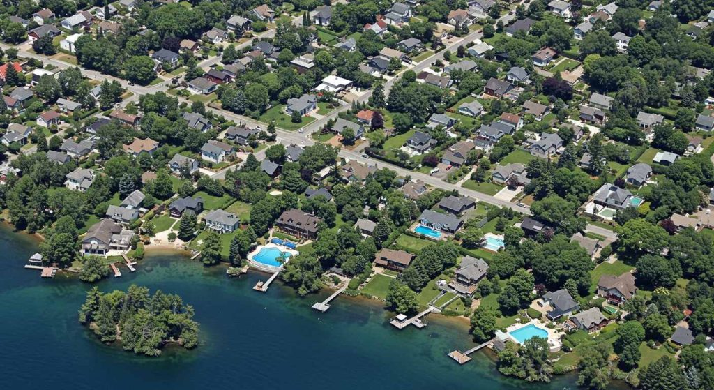 Aerial view of a suburban neighborhood in Clear Lake, Iowa, showcasing houses nestled among green trees, inviting swimming pools, and peaceful waterfront docks.