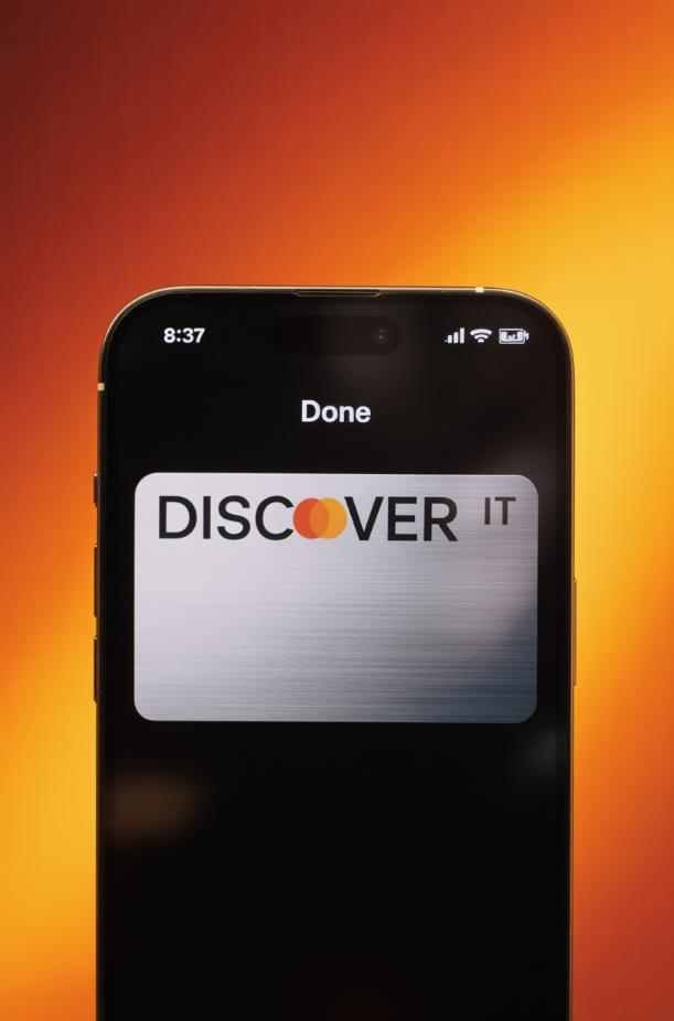 A smartphone displays a digital Discover It card against an orange gradient background, appealing to homeowners interested in Real Estate Investment Books or those looking to buy and flip properties.