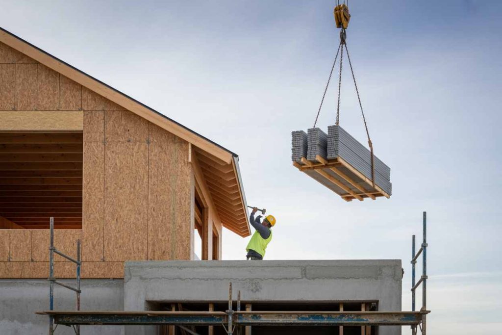 Construction worker hammers on a building as a crane lifts metal sheets beside a partially built house—modern methods like credit card alternatives are shaping how interior design businesses operate in 2025.