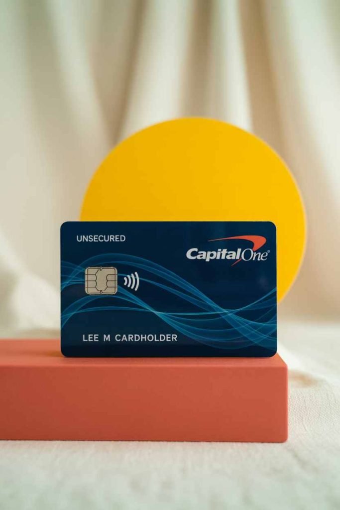 A Capital One credit card rests upright on a coral block with a yellow circle in the background—perfect for those looking to fund a home renovation or kickstart their DIY credit repair journey.