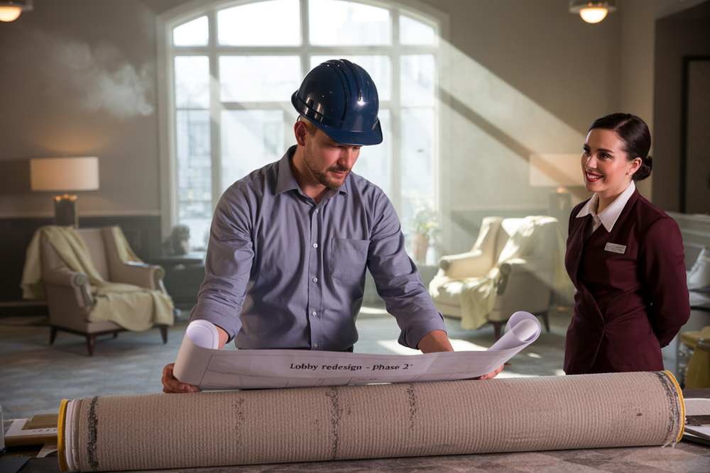 Man in hard hat reviews blueprints while woman in uniform smiles nearby in a well-lit room, reflecting the teamwork often seen during hotel renovation projects.