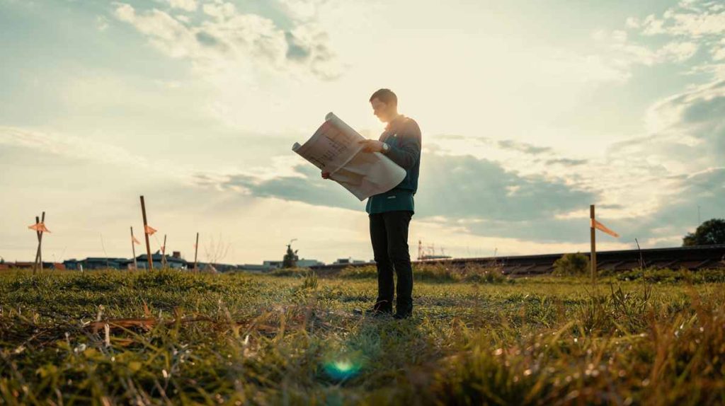 Person standing in a grassy field at sunset, holding and examining large blueprints for a sustainable eco-home under a cloudy sky.