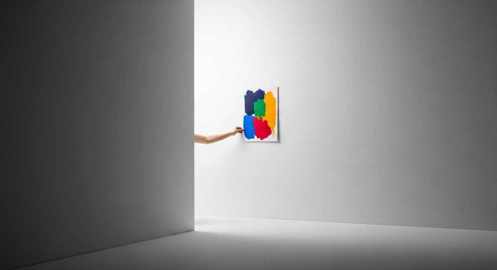A hand hangs a colorful abstract painting on a white wall, partially hidden by a large gray partition—a quiet moment in the architecture of settling in.