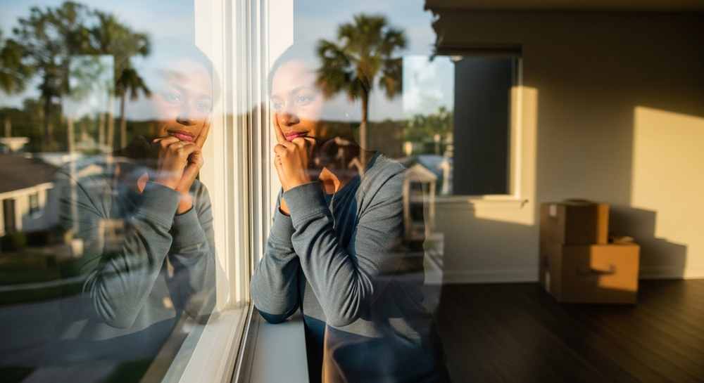 Woman looks thoughtfully out a window, reflected in the glass; unpacked boxes and sunlight hint at her big move and the early days of settling in.