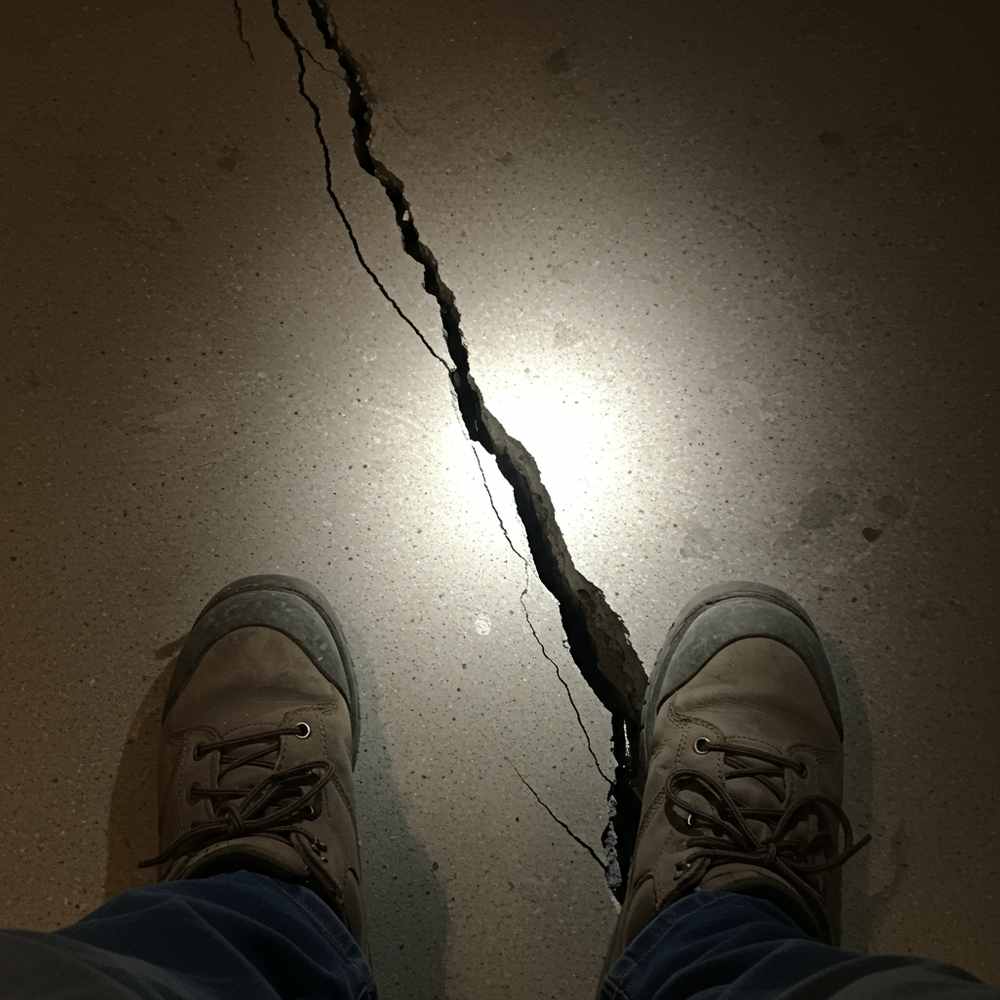 A pair of boots near a large crack running through a concrete floor, illuminated by a flashlight, suggests a possible foundation problem.